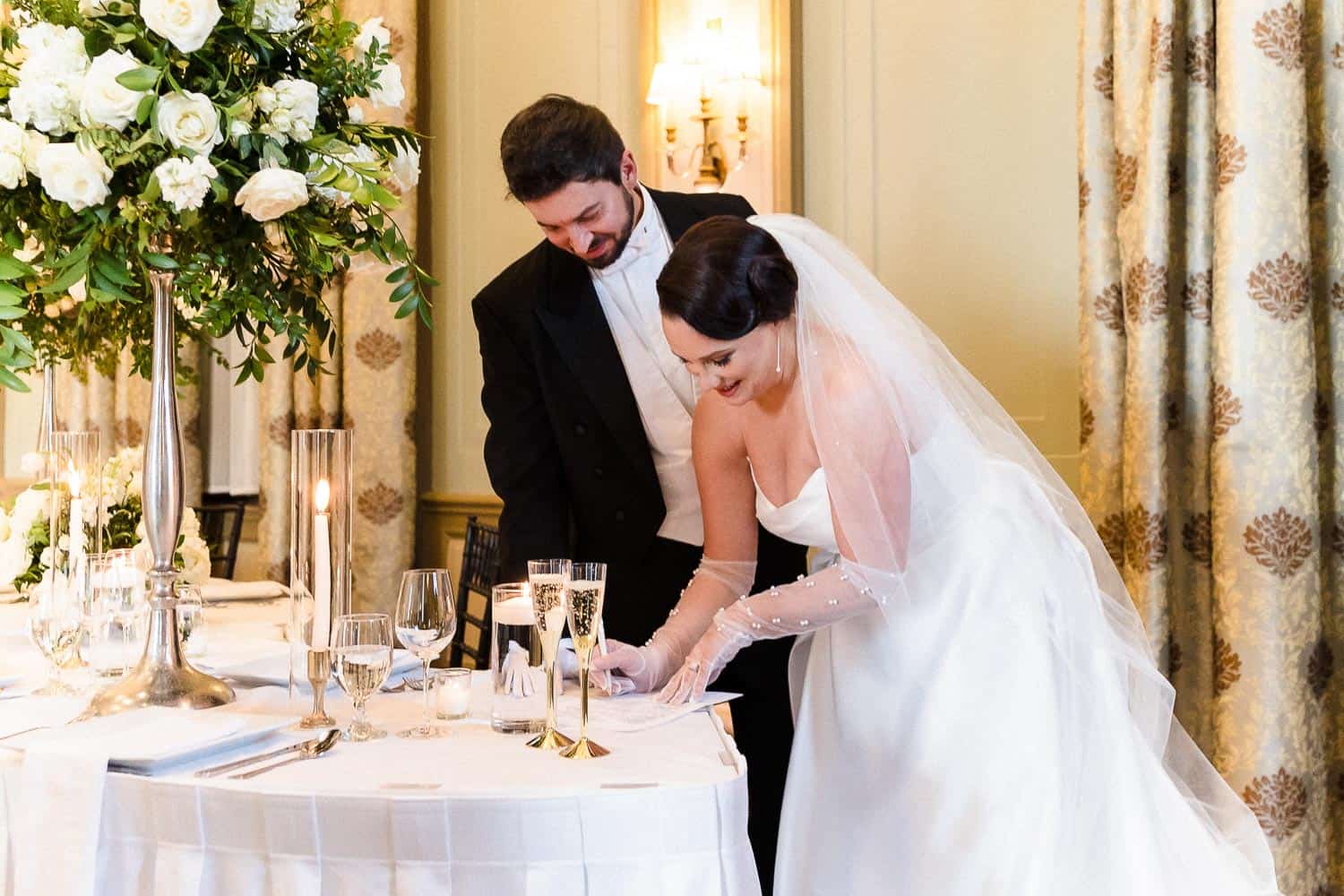 A bride and groom sign a document at a decorated table during their wedding ceremony, with flower arrangements and lit candles in the background.