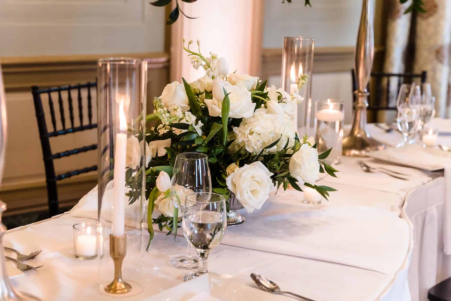 Elegant table setting with a white floral centerpiece, candles, glassware, and cutlery on a white tablecloth. Black chairs are visible in the background.