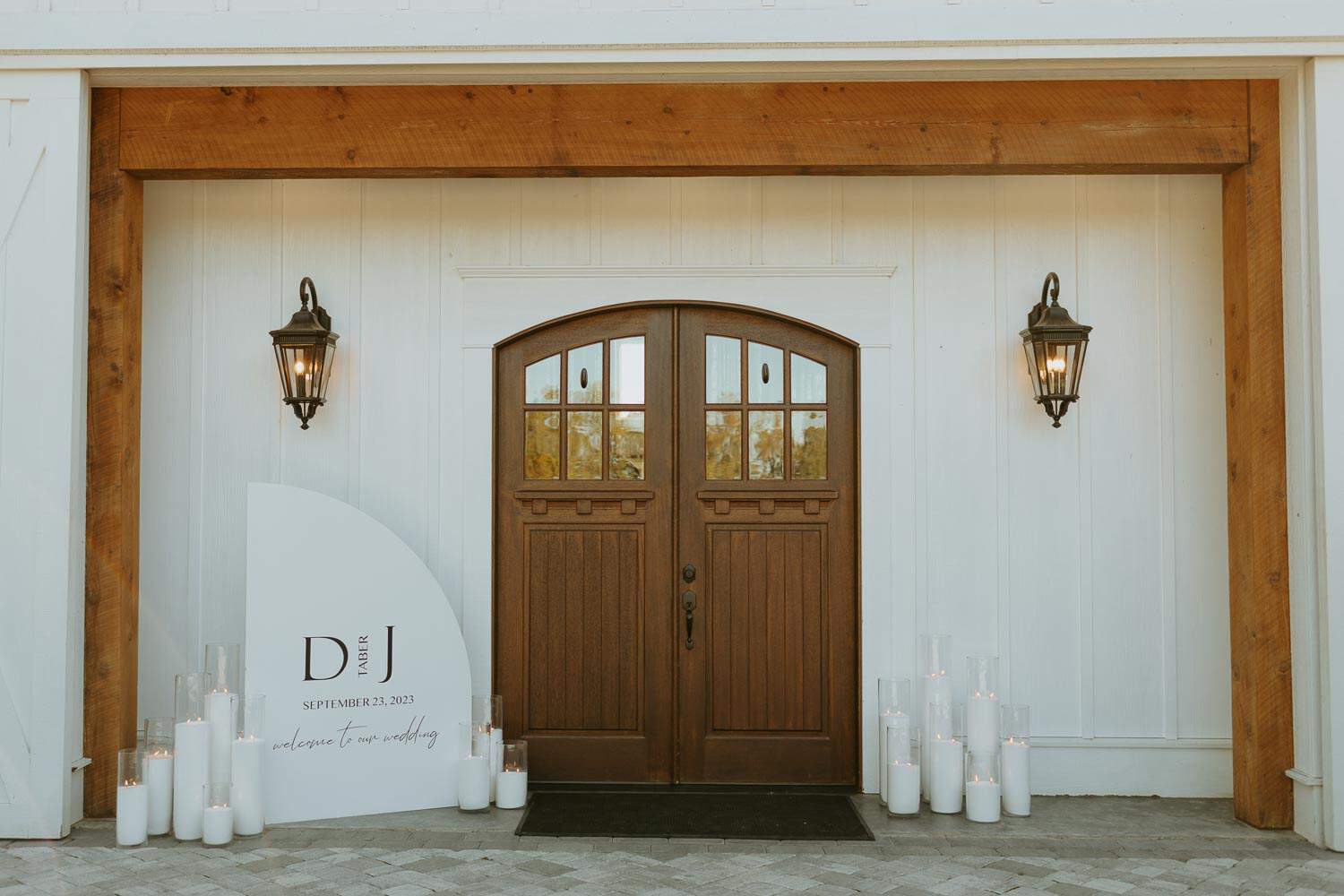 Wooden double doors at a white building, framed by two lanterns. A decorative sign with initials "D and J" plus a date and candles sit on the ground below.