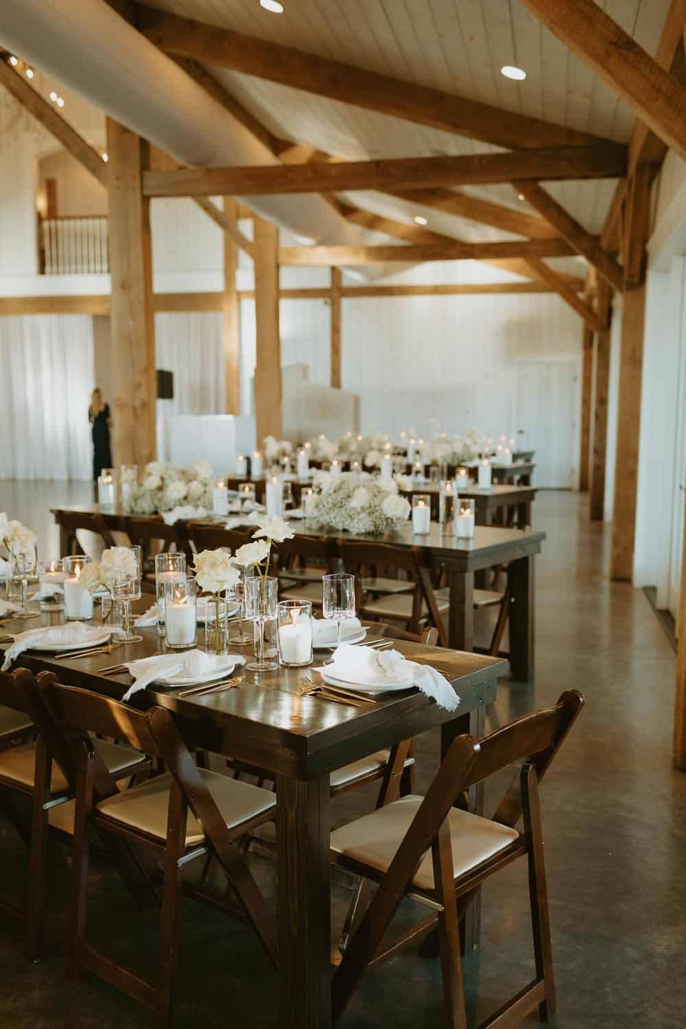 Spacious rustic venue with wooden beams and long tables set with white flowers, candles, folded napkins, and glassware.