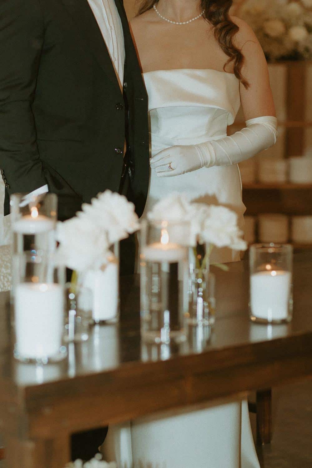 Bride in a strapless gown with gloves and groom in a dark suit stand together. Candles and flowers in glass vases are on a table in the foreground.