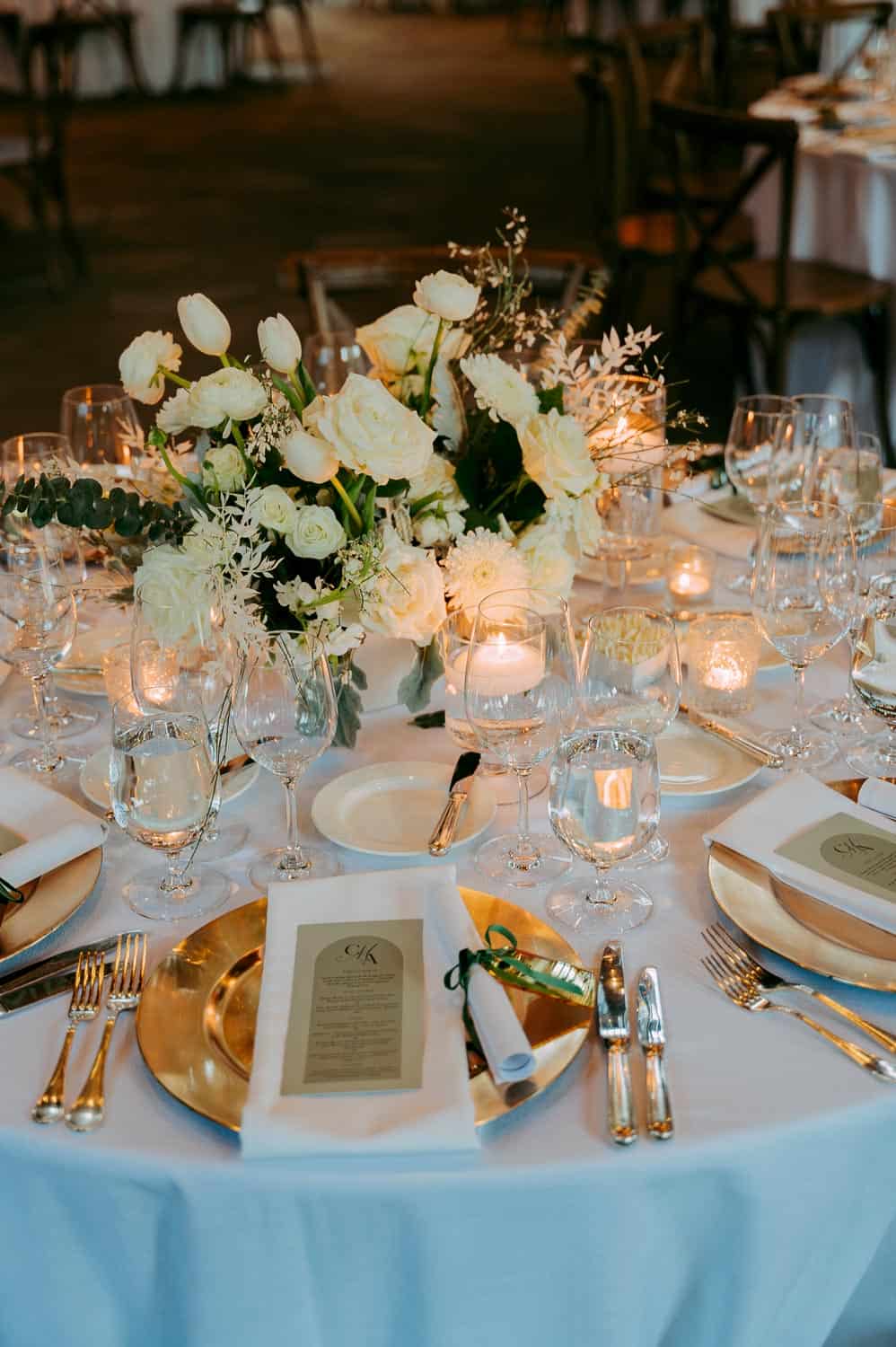 Elegant table setting with white floral centerpiece, gold plates, menus, and glassware arranged around lit candles on a round table.