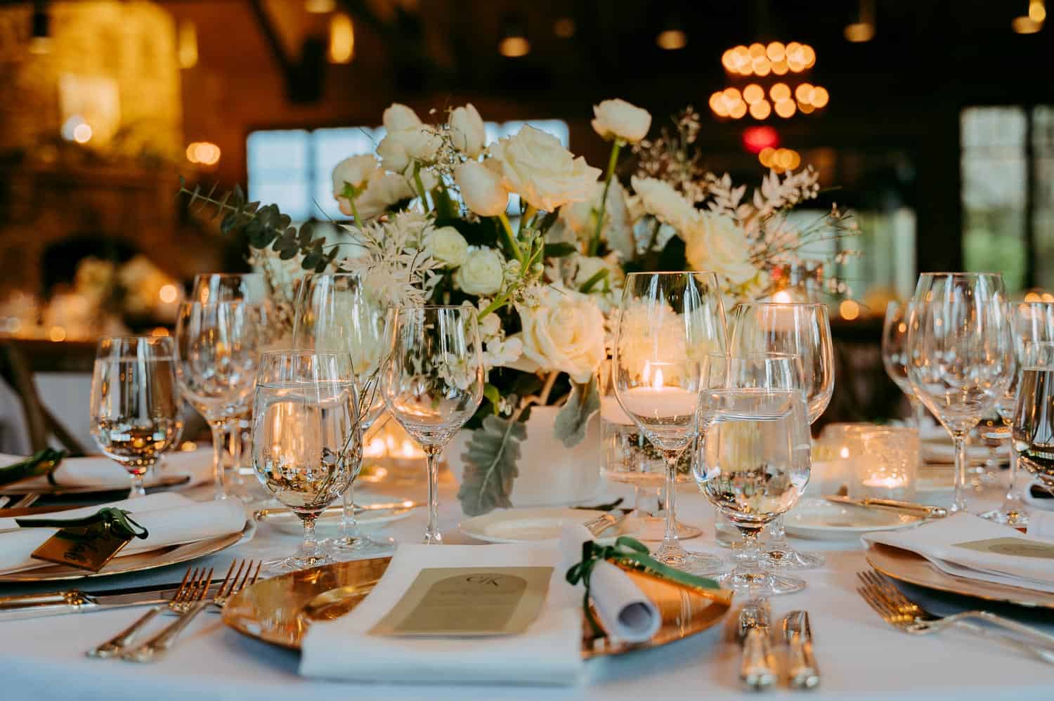 Elegant table setting with white flowers, candles, and glassware on a round table with a white tablecloth, set for a formal event in a warmly lit indoor venue.