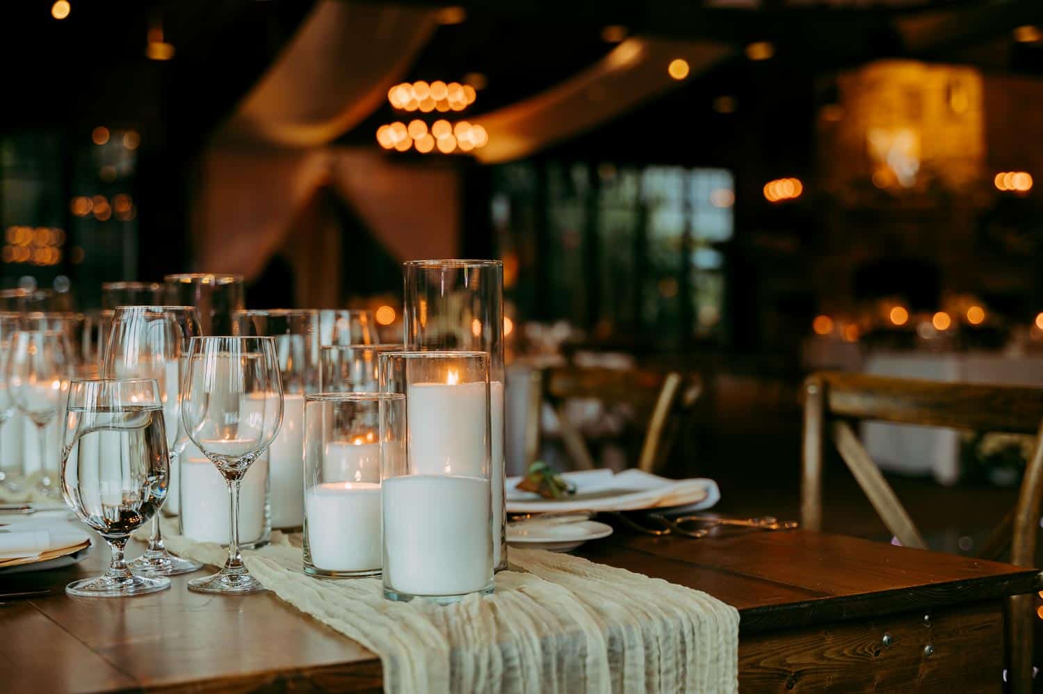 A wooden table with lit candles in glass holders, wine glasses, and a decorative cloth runner. The background features soft lighting and draped fabric.