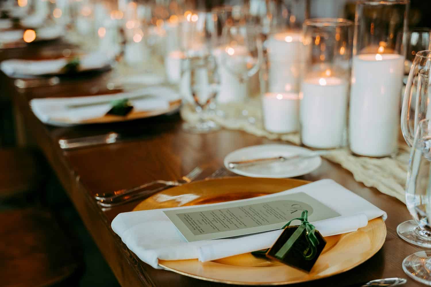 Elegant table setting with a menu and gift box on each plate, white candles and glassware arranged in a row on a wooden table.