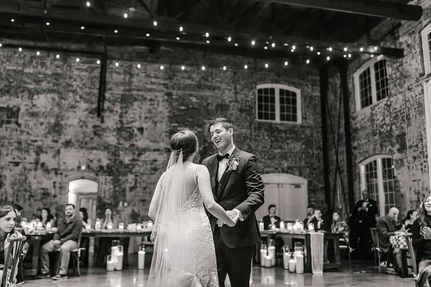 A couple in wedding attire is dancing in a rustic venue with exposed brick walls and string lights, while guests seated at tables watch.