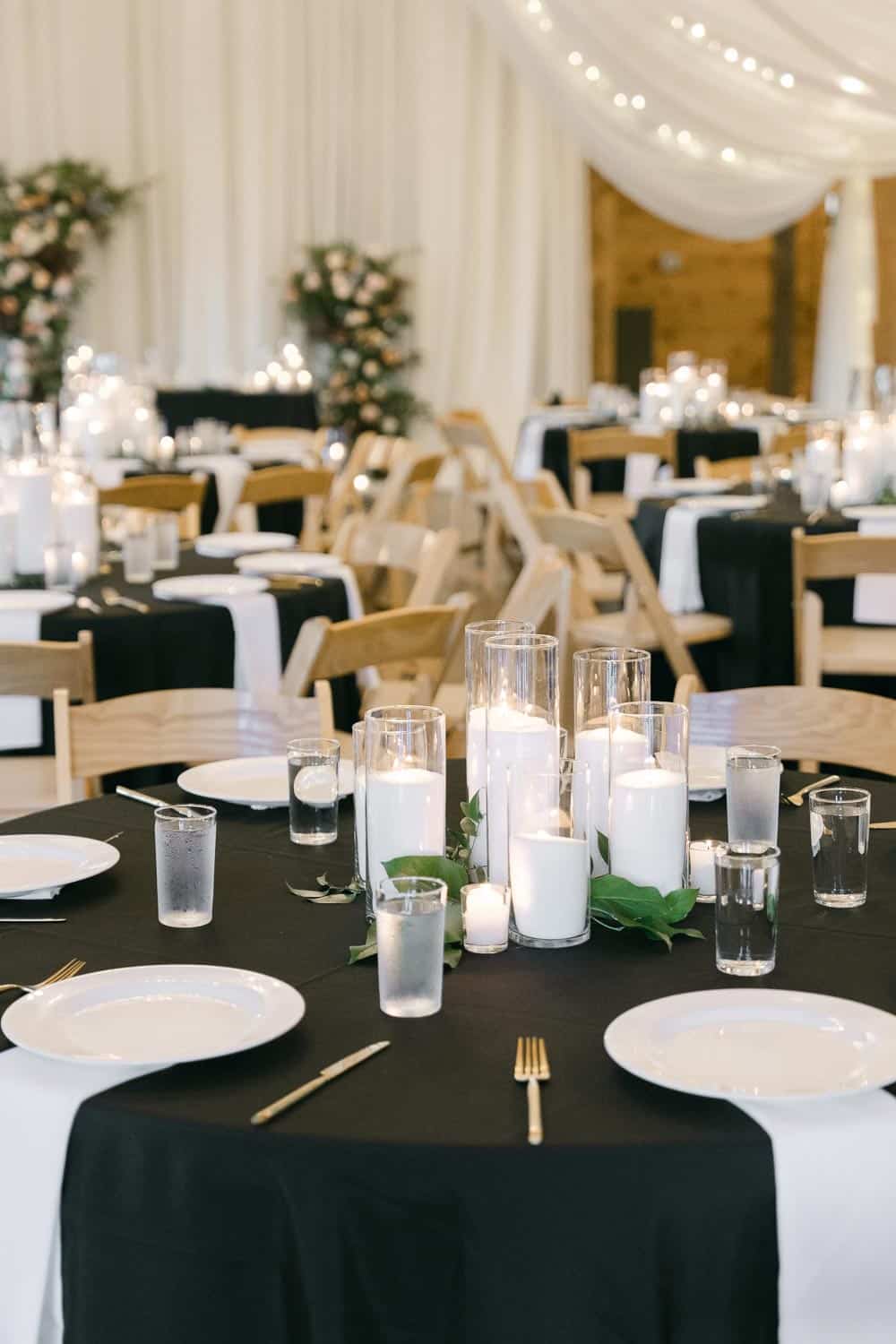 Wedding reception with round tables in black and white tablecloths, set with candles, glasses, and plates. Wooden chairs surround the tables.