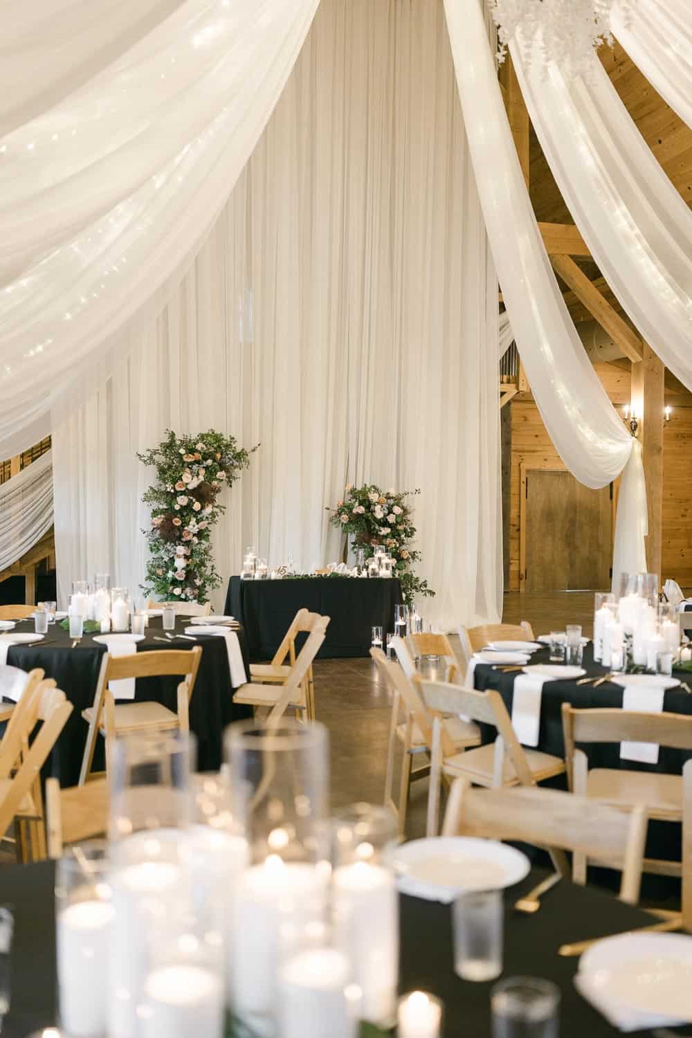 A decorated wedding reception hall with draped white fabric, round tables with black tablecloths, white chairs, candles, and floral arrangements.