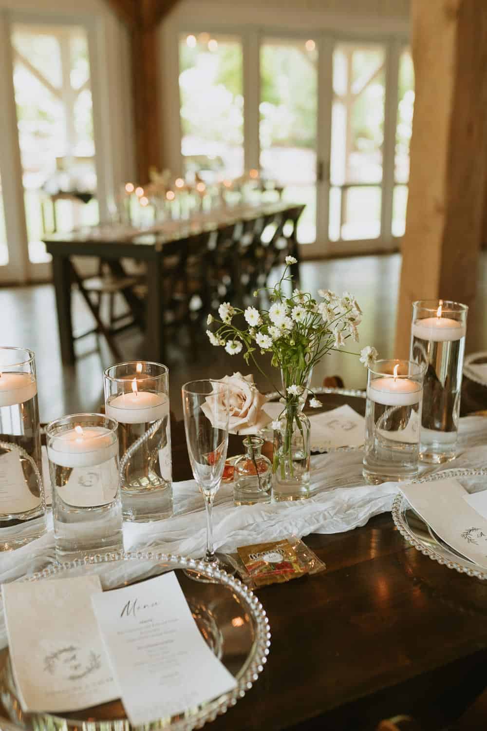 Elegant table with floating candles, a floral arrangement, and menu cards on clear chargers. Background shows tables and large windows with greenery.