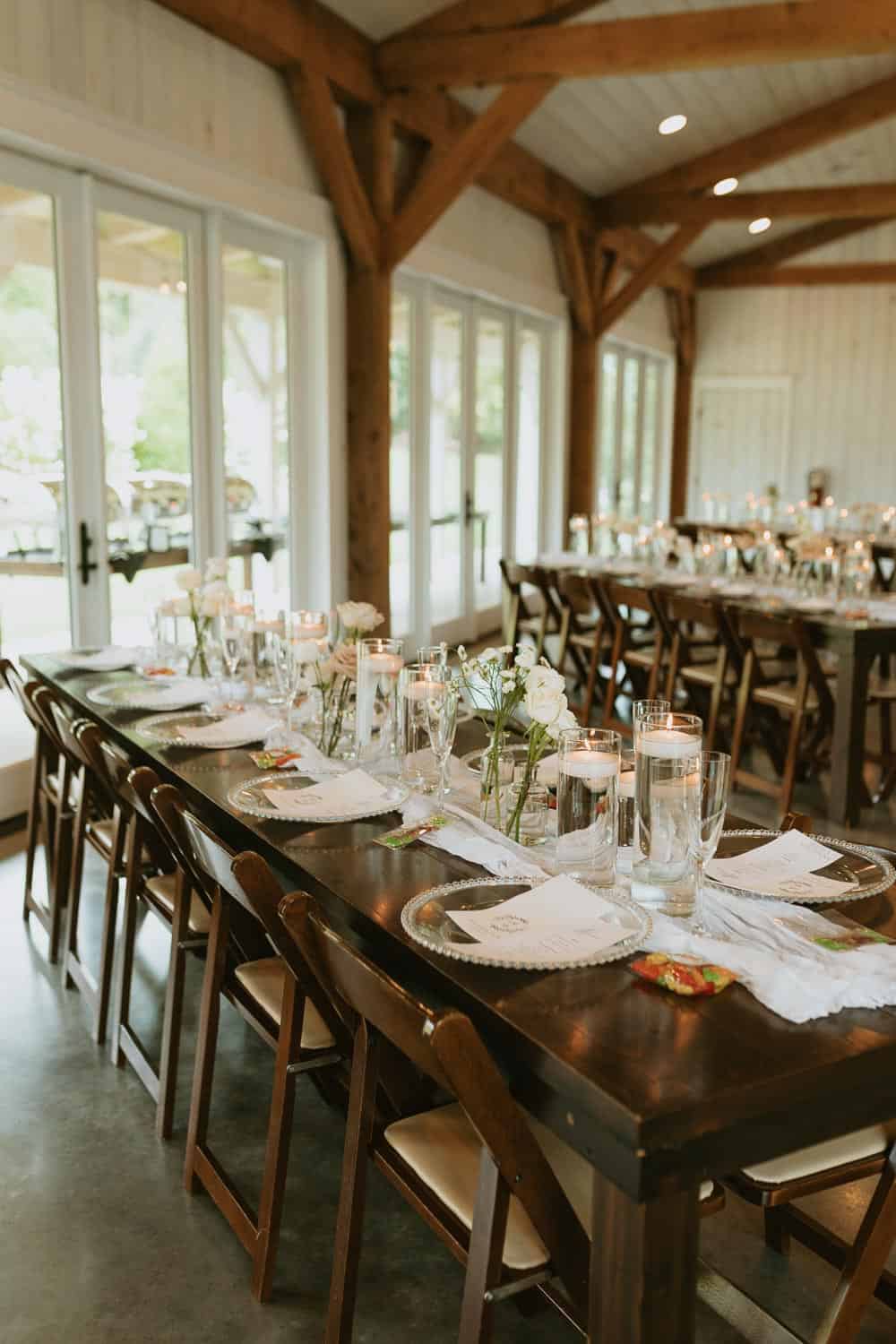 A rustic wooden table set for a formal gathering with plates, glasses, and white floral centerpieces in a bright room with large windows and wooden beams.