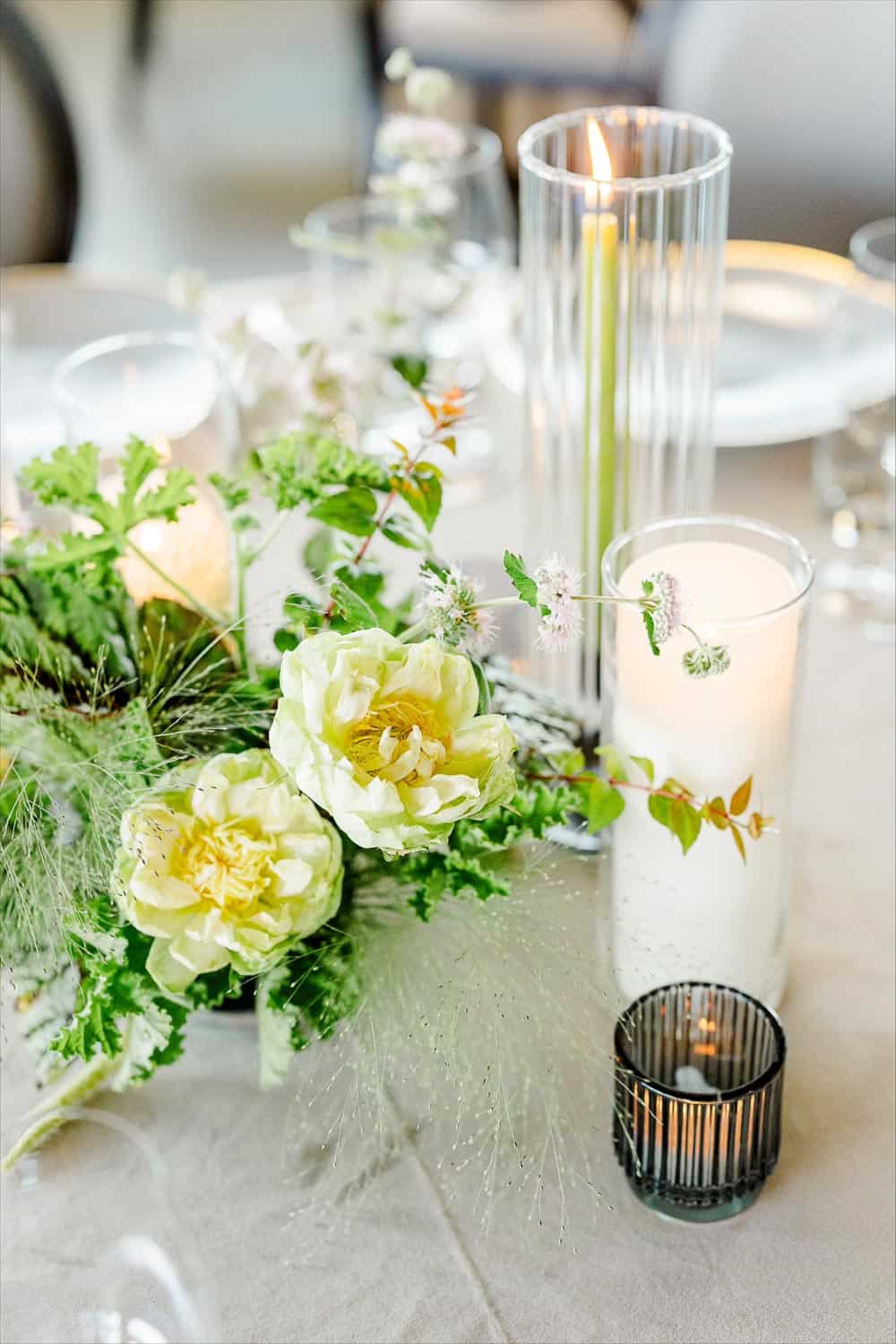 A tabletop display with white flowers, green foliage, and three candles in glass holders on a beige tablecloth.