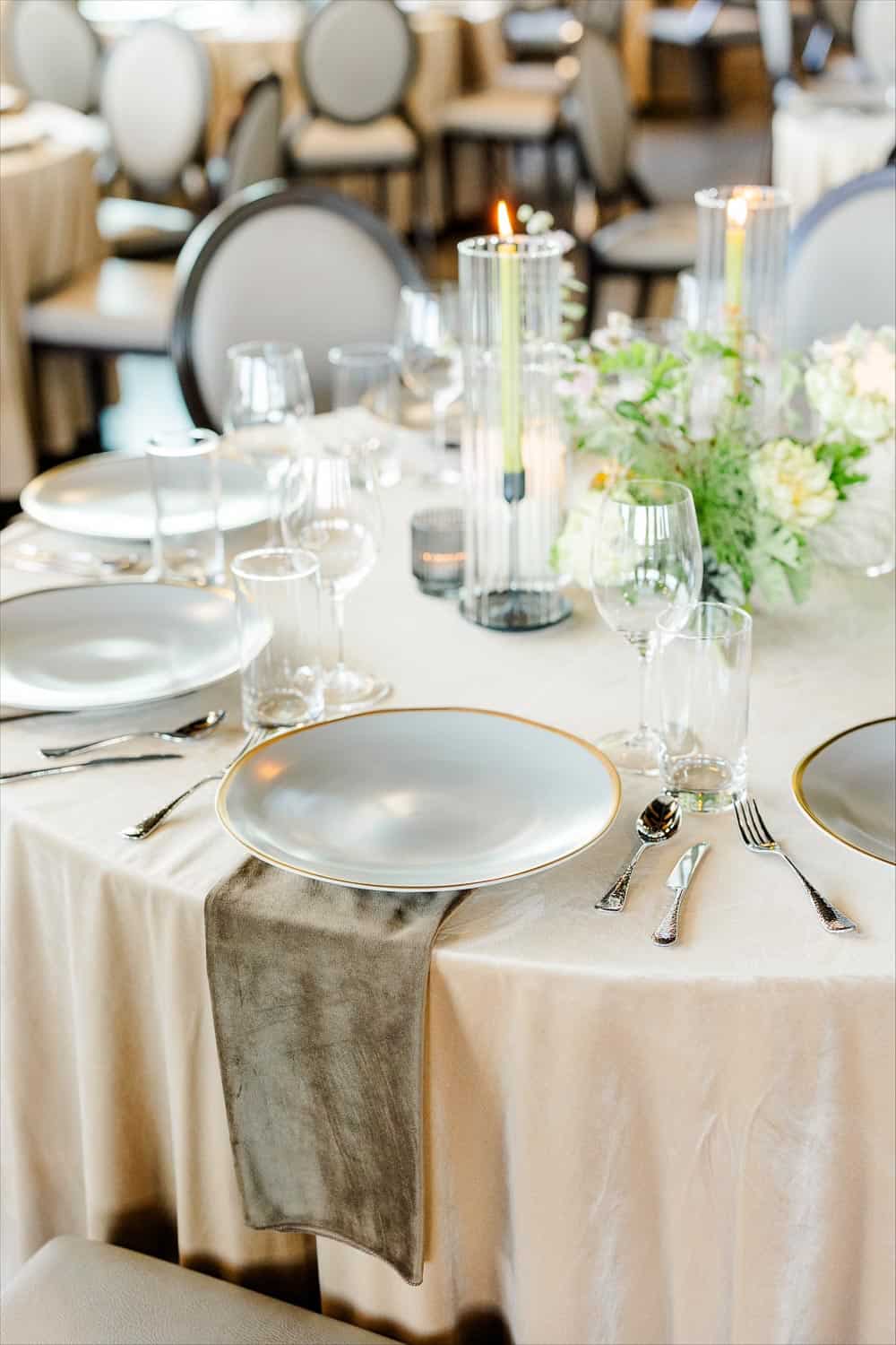 Elegant table setting with silver plates, cutlery, glassware, gray napkins, and floral centerpiece on a draped table.