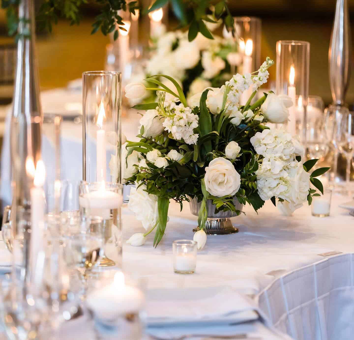 Elegant table setting with white floral centerpieces, tall candles in glass holders, and wine glasses on a white tablecloth.