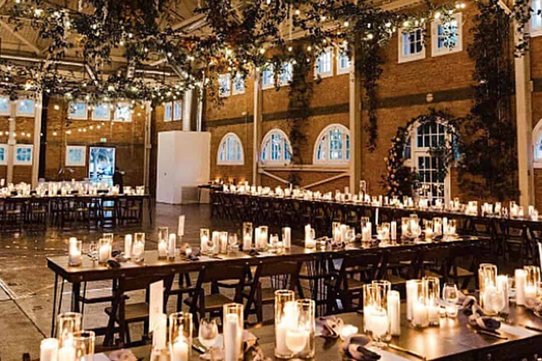 A warmly lit banquet hall with long tables set up for an event. Candles line the tables, and string lights hang from the ceiling.
