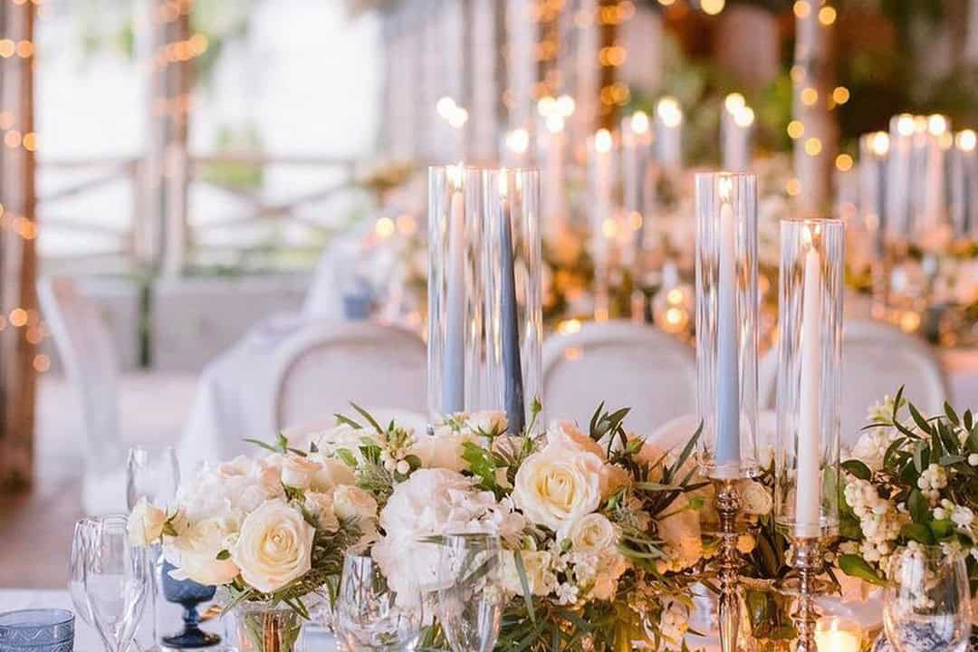 Elegant table setting with white roses, greenery, and glass candleholders. Soft lighting and blurred background create a warm ambiance.
