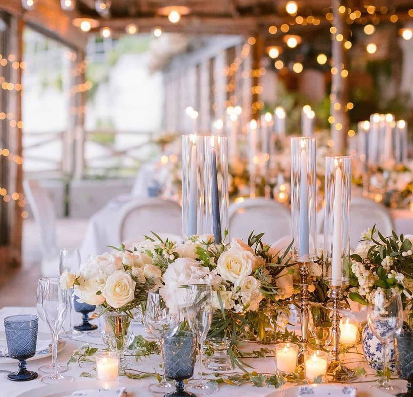 Elegant table setting with white roses, candles, and glassware. Soft lighting and string lights create a warm ambiance.