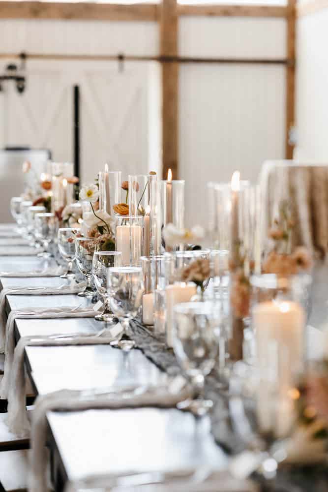 Elegant dining table set with white napkins, glassware, and floral arrangements, featuring lit candles in glass holders, in a bright room with wooden accents.