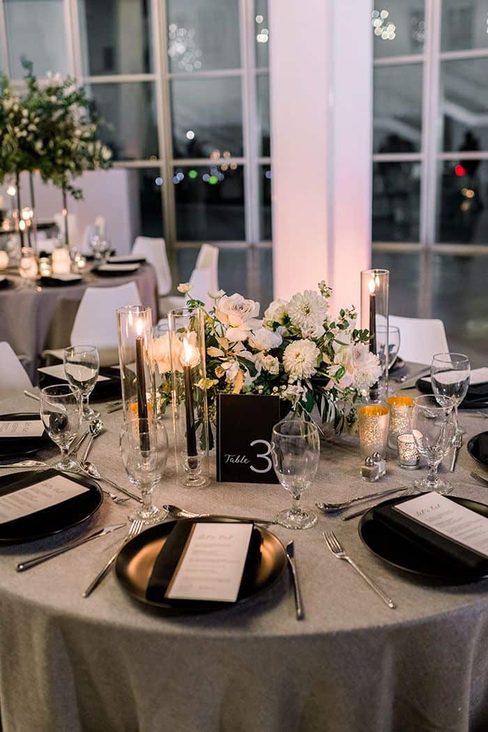 Elegant table setting with black plates, silverware, glasses, and a floral centerpiece of white flowers and candles on a gray tablecloth. "Table 3" card visible.