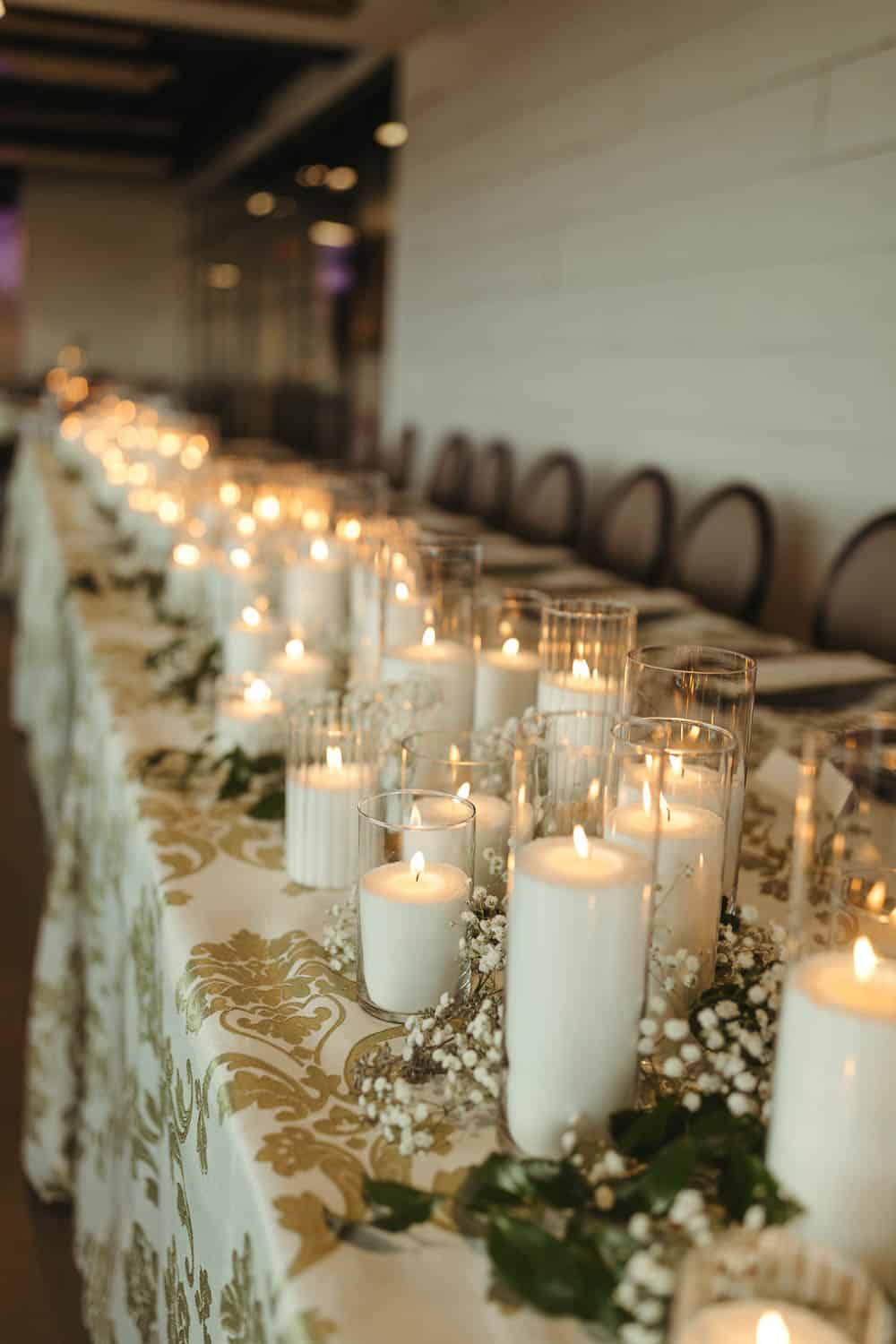 Long table with lit candles in glass holders, greenery, a patterned tablecloth, and chairs arranged on one side.