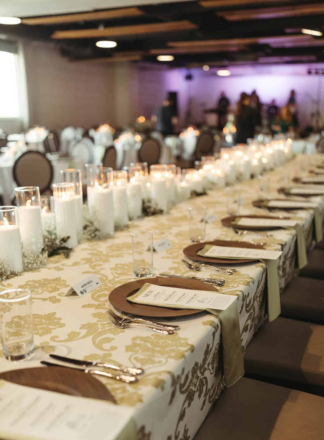 Elegant table setting with floral tablecloth, place settings, and candles in clear holders. Chairs are arranged around the table in a softly lit room.