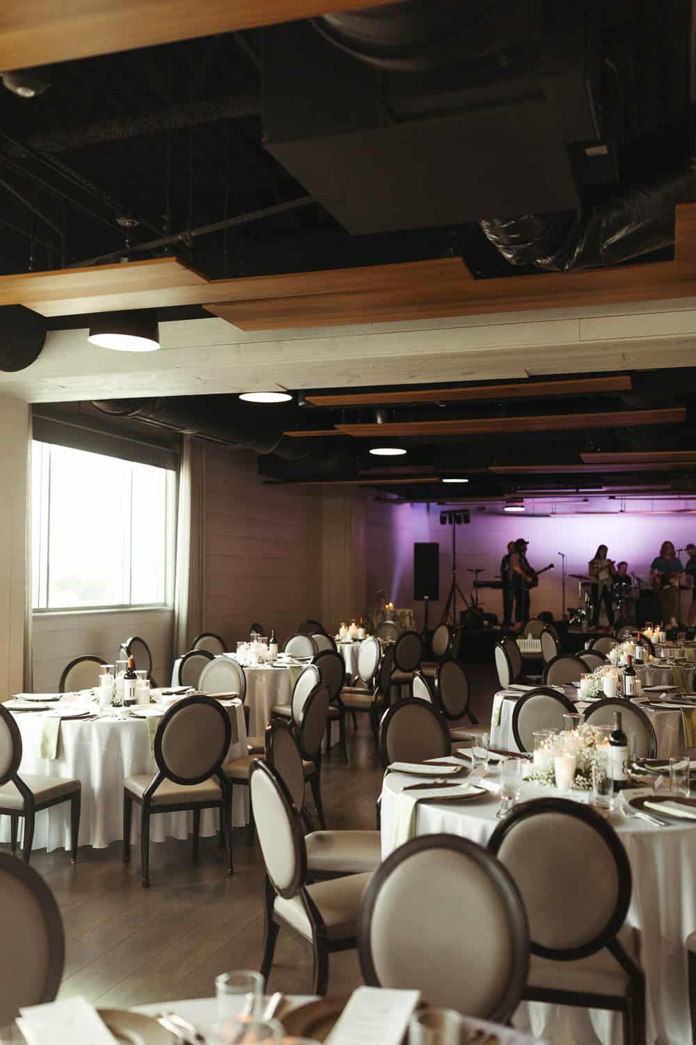 A room with round tables set for a banquet, featuring white tablecloths and chairs. A band sets up on a stage in the background, with purple lighting overhead.