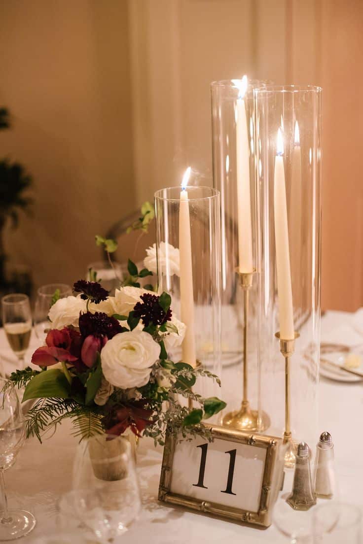 Table setting with tall candles in glass holders, a floral arrangement with white and red flowers, and a table number 11 card.