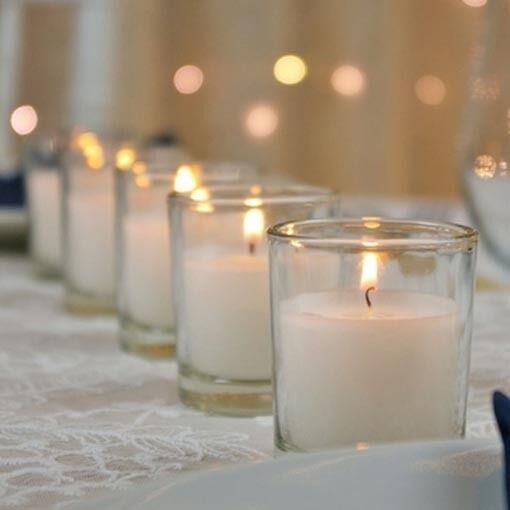 A row of lit white candles in glass holders on a lace-covered table, with softly blurred lights in the background.