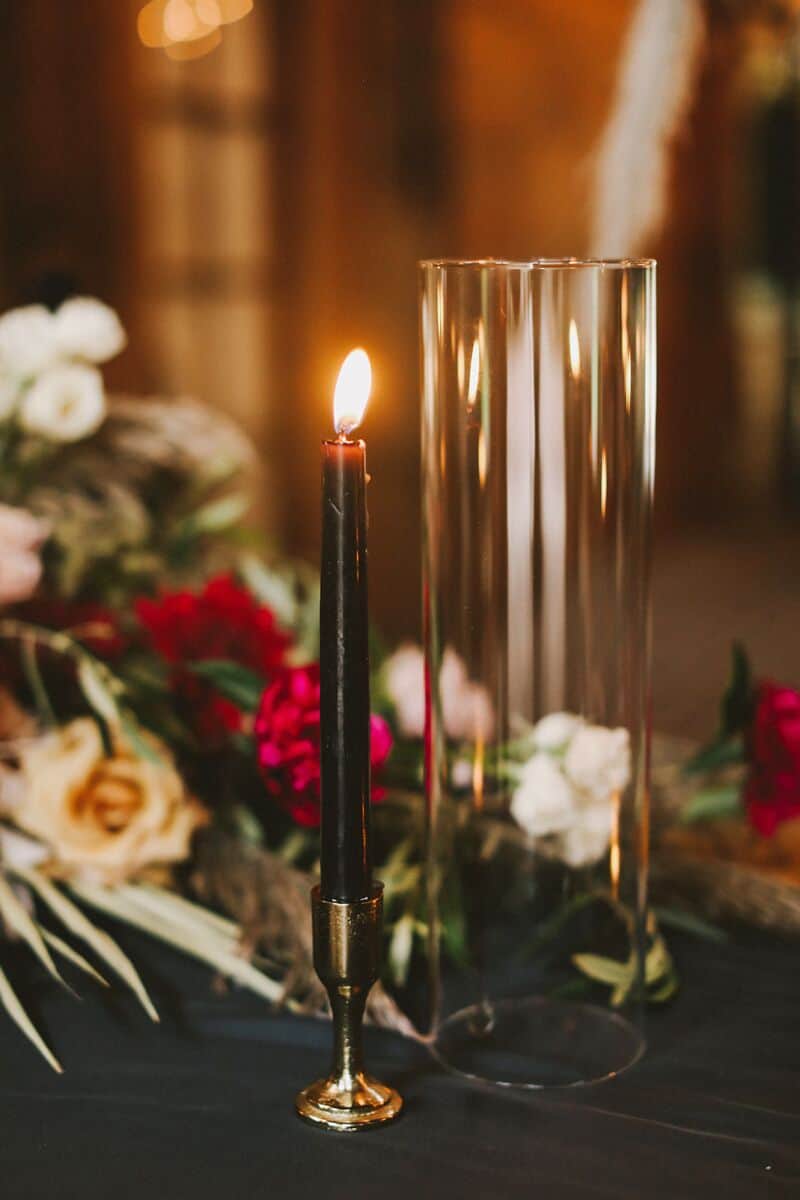 A lit black candle in a brass holder next to a transparent glass vase, with red and white flowers in the background.