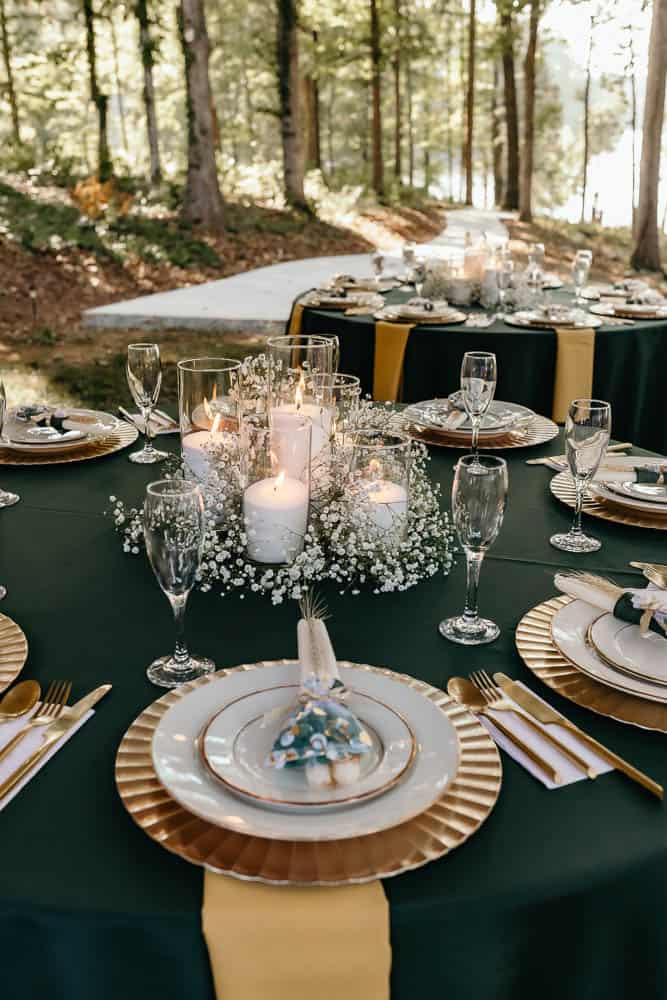 Elegant outdoor table with green and gold decor, lit candles in glass holders, white plates on gold chargers, champagne flutes, and a baby’s breath centerpiece.