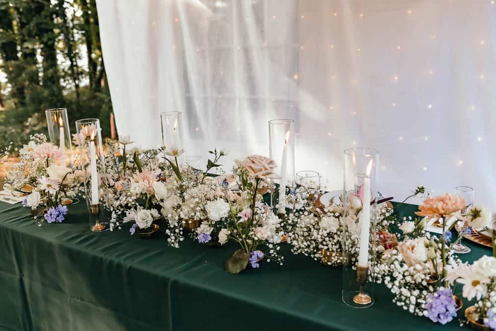A table centerpiece with white and pink flowers, greenery, and lit tall candles is set on a green tablecloth. A twinkling backdrop is visible in the background.