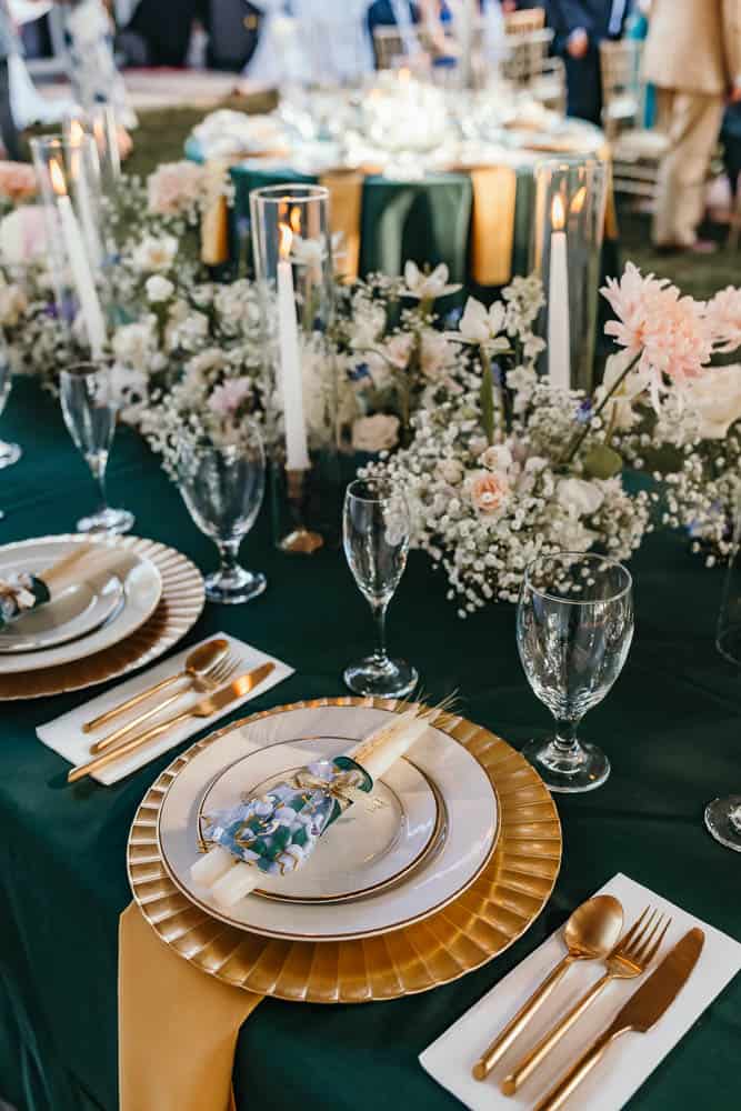 Elegant table setting with gold and white plates, gold cutlery, and white napkins. Floral centerpieces with candles decorate the table.
