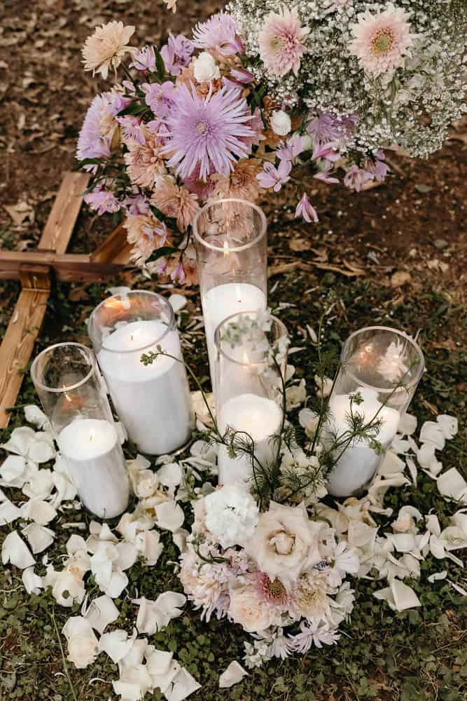Candles in glass holders surrounded by white rose petals and floral arrangements, next to a wooden cross on the ground.