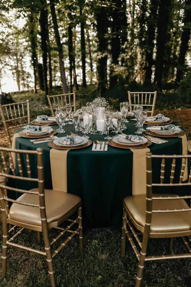 Outdoor dining setup with a round table covered in a dark green tablecloth, set with plates, glasses, and cutlery. Surrounded by six gold chairs, in a wooded area.