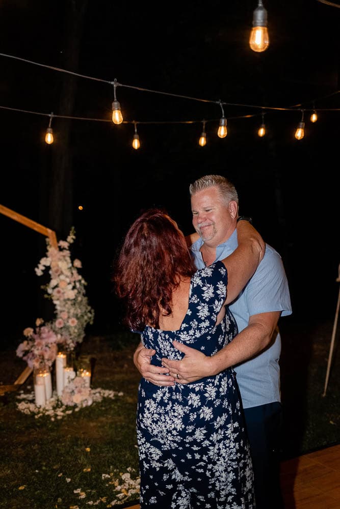 A couple dances together outside under string lights at night, surrounded by flowers and a small arch.